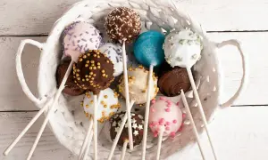 Kosz pełen kolorowych cake popsów: czekoladowych, białych, różowych i niebieskich, ozdobionych posypkami i złotymi kuleczkami.