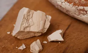 Kostka świeżych drożdży, pokruszona na drewnianej desce, obok bochenka chleba. Tak wyglądają drożdże przed pieczeniem.