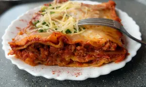 Pyszna lasagne bolognese z warstwami makaronu, mięsa mielonego i sosu pomidorowego, posypana serem i natką pietruszki. Widelec czeka na pierwszy kęs.