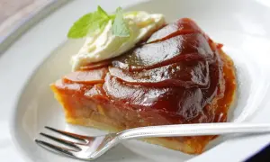 Kawałek karmelizowanej tarta tatin z jabłkami, podany z bitą śmietaną i miętą, na białym talerzu z widelcem.