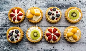 Osiem kolorowych tartaletek z owocami: truskawkami, borówkami, kiwi, bananami i morelami, na kremowym nadzieniu.