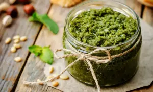 Słoik domowego pesto bazyliowego, ozdobiony sznurkiem. Obok leżą orzeszki piniowe i listki bazylii.