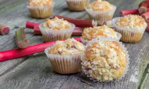 Pyszne muffinki z rabarbarem, posypane kruszonką, leżą na drewnianym blacie obok świeżych łodyg rabarbaru.