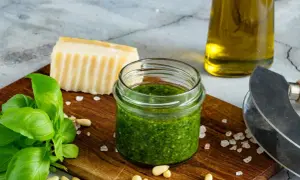 Słoiczek domowego pesto z bazylii, obok świeże liście bazylii, orzeszki piniowe, kawałek parmezanu i oliwa.