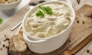 Aksamitny sos pieczarkowy w białej miseczce, udekorowany natką pietruszki. Obok świeże pieczarki i ziarna pieprzu.