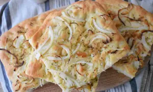 Puszysta focaccia z cebulą, posypana ziołami i solą, prezentuje się apetycznie na drewnianej desce.