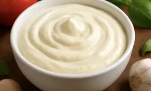 Gładki, kremowy sos aioli w białej miseczce, obok czosnek, pomidor i bazylia.