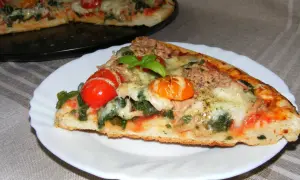 Kawałek pysznej pizzy z tuńczykiem, pomidorkami i bazylią na talerzyku. W tle cała pizza.