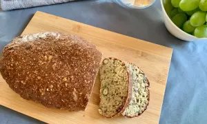 Chleb z twarogu, bogaty w ziarna i orzechy, pokrojony na desce. Obok miska z winogronami i szklanka wody.