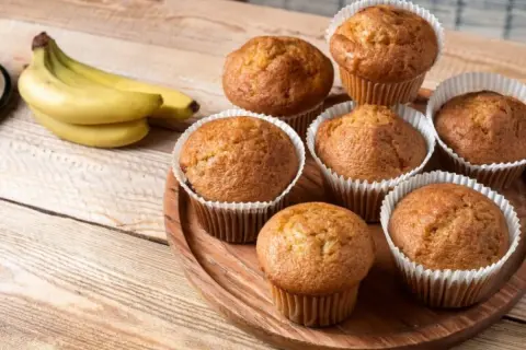 Puszyste babeczki bananowe, świeżo upieczone, kuszą swoim aromatem. Obok leży kiść dojrzałych bananów.