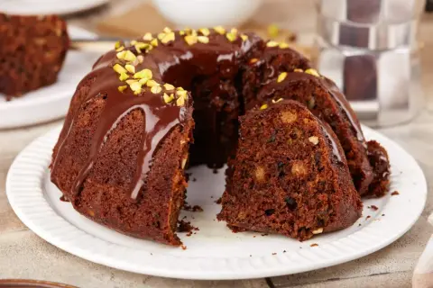 Pyszna babka na oleju, polana czekoladą i posypana pistacjami, z kawałkiem ciasta obok.