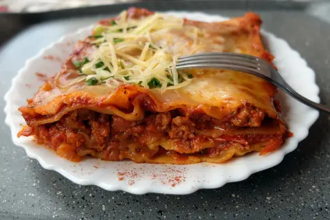 Pyszna lasagne bolognese z warstwami makaronu, mięsa mielonego i sosu pomidorowego, posypana serem i natką pietruszki. Widelec czeka na pierwszy kęs.