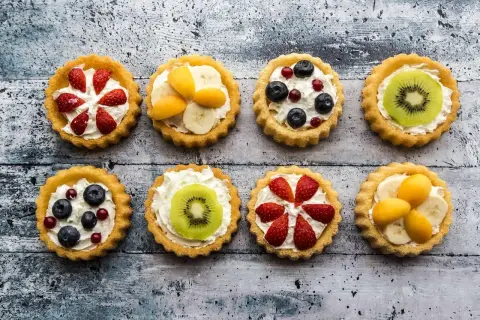 Osiem kolorowych tartaletek z owocami: truskawkami, borówkami, kiwi, bananami i morelami, na kremowym nadzieniu.
