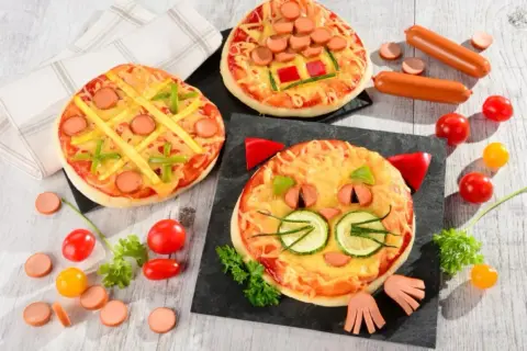 Wesołe pizza dla dzieci: jedna w kształcie kota, druga jak kółko i krzyżyk, trzecia z parówkami i papryką.