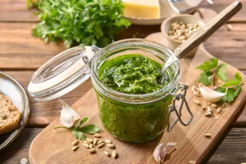 Słoik domowego pesto z pietruszki, obok czosnek, nasiona słonecznika i kawałek chleba.