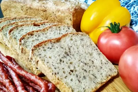 Pyszny chleb nocny na zakwasie, pokrojony w kromki, obok wędzonej kiełbasy, pomidorów i żółtej papryki.
