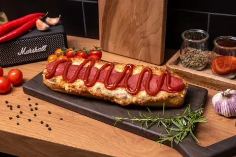 Pyszna zapiekanka z salami, posypana serem i polana ketchupem, prezentuje się apetycznie na desce. Obok czosnek, pomidorki i przyprawy.