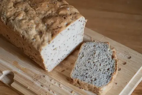 Chleb z siemienia lnianego, z jedną kromką obok, na desce do krojenia.