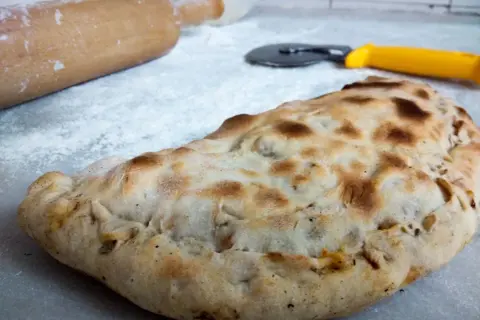 Złocisty calzone, gotowy do podania. Obok wałek i nóż do pizzy, jakby ktoś właśnie skończył przygotowywać ten pyszny przepis.