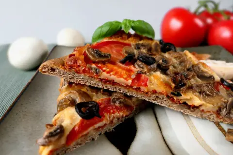 Dwie kromki pizzy na cieście z mąki pełnoziarnistej z pieczarkami, oliwkami i pomidorami.