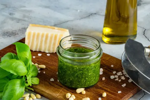 Słoiczek domowego pesto z bazylii, obok świeże liście bazylii, orzeszki piniowe, kawałek parmezanu i oliwa.