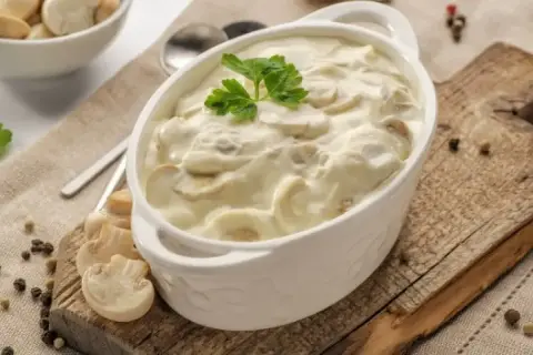 Aksamitny sos pieczarkowy w białej miseczce, udekorowany natką pietruszki. Obok świeże pieczarki i ziarna pieprzu.