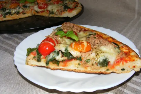 Kawałek pysznej pizzy z tuńczykiem, pomidorkami i bazylią na talerzyku. W tle cała pizza.