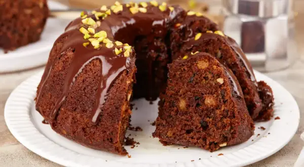 Pyszna babka na oleju, polana czekoladą i posypana pistacjami, z kawałkiem ciasta obok.