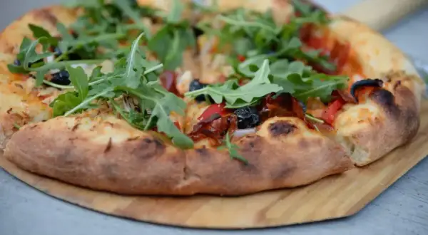 Pyszne ciasto na pizzę z rukolą, oliwkami i papryką, podane na drewnianej desce.