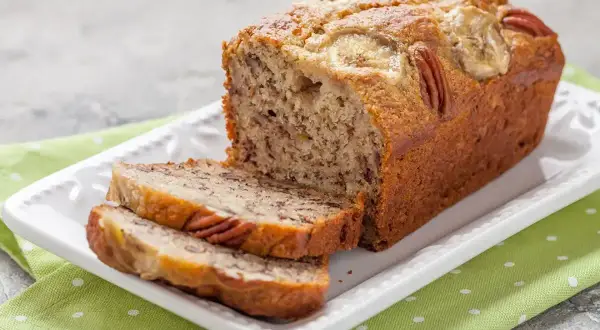 Pyszny chlebek bananowy z orzechami pekan i plasterkami banana na wierzchu, pokrojony na kromki, na białej paterze.