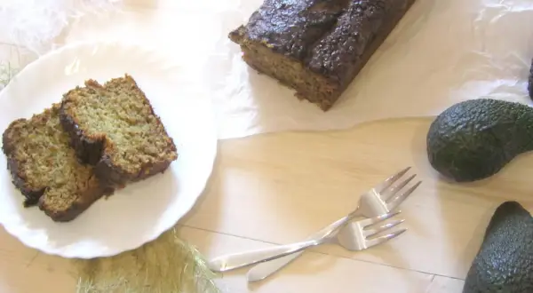 Chleb bananowy z awokado, pokrojony na kromki, leży na talerzu obok całego bochenka. Dwa awokado i widelce dopełniają kompozycję.