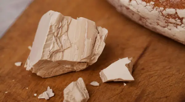 Kostka świeżych drożdży, pokruszona na drewnianej desce, obok bochenka chleba. Tak wyglądają drożdże przed pieczeniem.