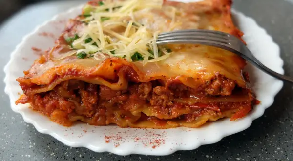 Pyszna lasagne bolognese z warstwami makaronu, mięsa mielonego i sosu pomidorowego, posypana serem i natką pietruszki. Widelec czeka na pierwszy kęs.