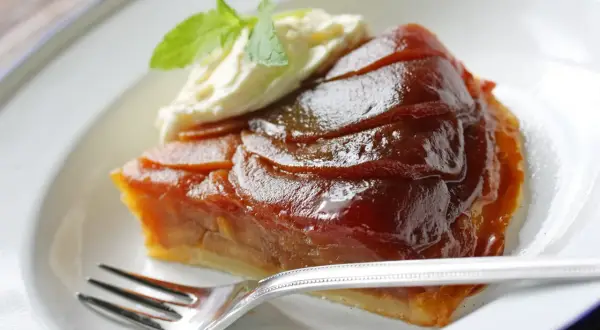 Kawałek karmelizowanej tarta tatin z jabłkami, podany z bitą śmietaną i miętą, na białym talerzu z widelcem.