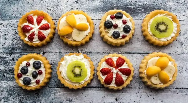 Osiem kolorowych tartaletek z owocami: truskawkami, borówkami, kiwi, bananami i morelami, na kremowym nadzieniu.
