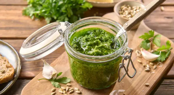 Słoik domowego pesto z pietruszki, obok czosnek, nasiona słonecznika i kawałek chleba.