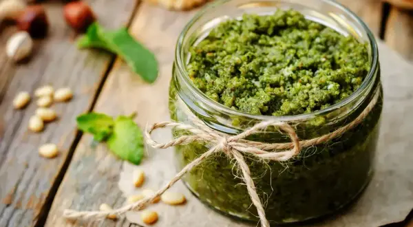 Słoik domowego pesto bazyliowego, ozdobiony sznurkiem. Obok leżą orzeszki piniowe i listki bazylii.