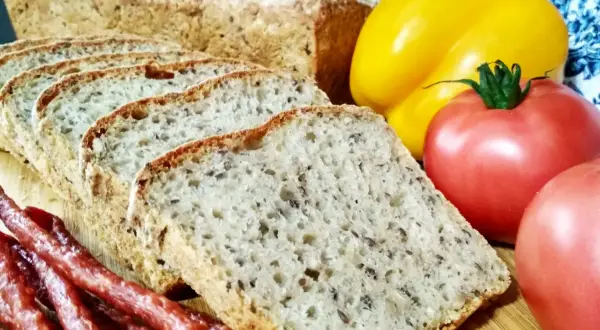Pyszny chleb nocny na zakwasie, pokrojony w kromki, obok wędzonej kiełbasy, pomidorów i żółtej papryki.