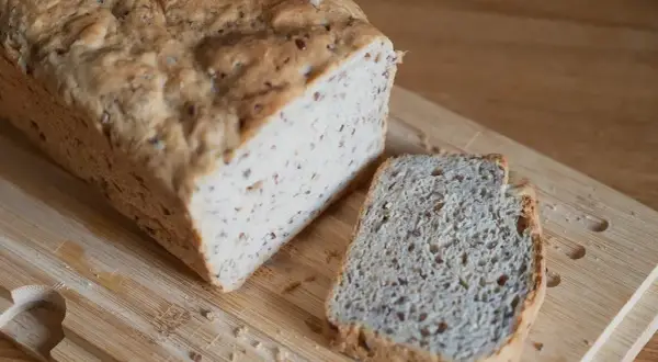 Chleb z siemienia lnianego, z jedną kromką obok, na desce do krojenia.