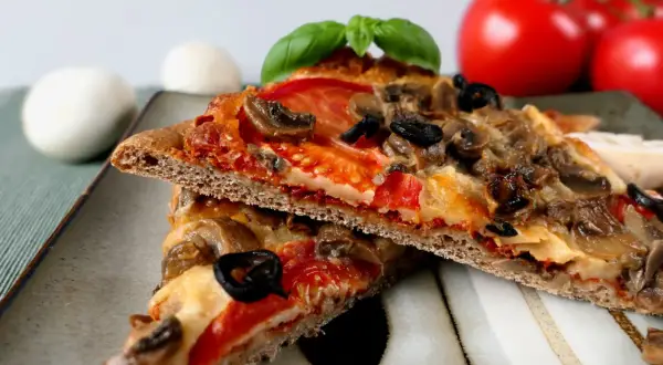 Dwie kromki pizzy na cieście z mąki pełnoziarnistej z pieczarkami, oliwkami i pomidorami.