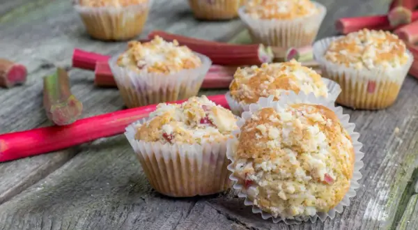 Pyszne muffinki z rabarbarem, posypane kruszonką, leżą na drewnianym blacie obok świeżych łodyg rabarbaru.