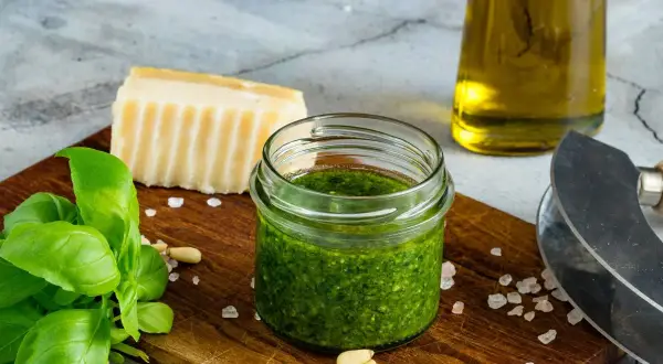 Słoiczek domowego pesto z bazylii, obok świeże liście bazylii, orzeszki piniowe, kawałek parmezanu i oliwa.