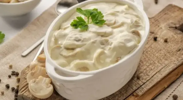 Aksamitny sos pieczarkowy w białej miseczce, udekorowany natką pietruszki. Obok świeże pieczarki i ziarna pieprzu.