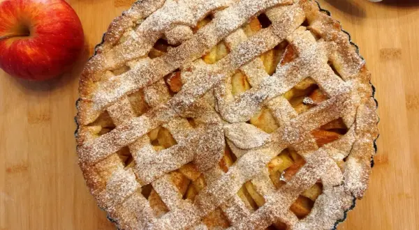 Pyszna tarta z jabłkami, posypana cukrem pudrem, czeka na podanie. Obok leżą świeże jabłka.