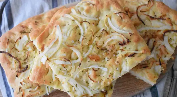 Puszysta focaccia z cebulą, posypana ziołami i solą, prezentuje się apetycznie na drewnianej desce.
