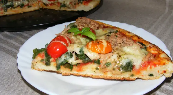 Kawałek pysznej pizzy z tuńczykiem, pomidorkami i bazylią na talerzyku. W tle cała pizza.
