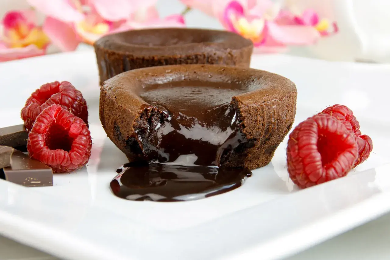 Płynny środek gorącego lava cake spływa na talerz, otoczony malinami i kawałkami czekolady.