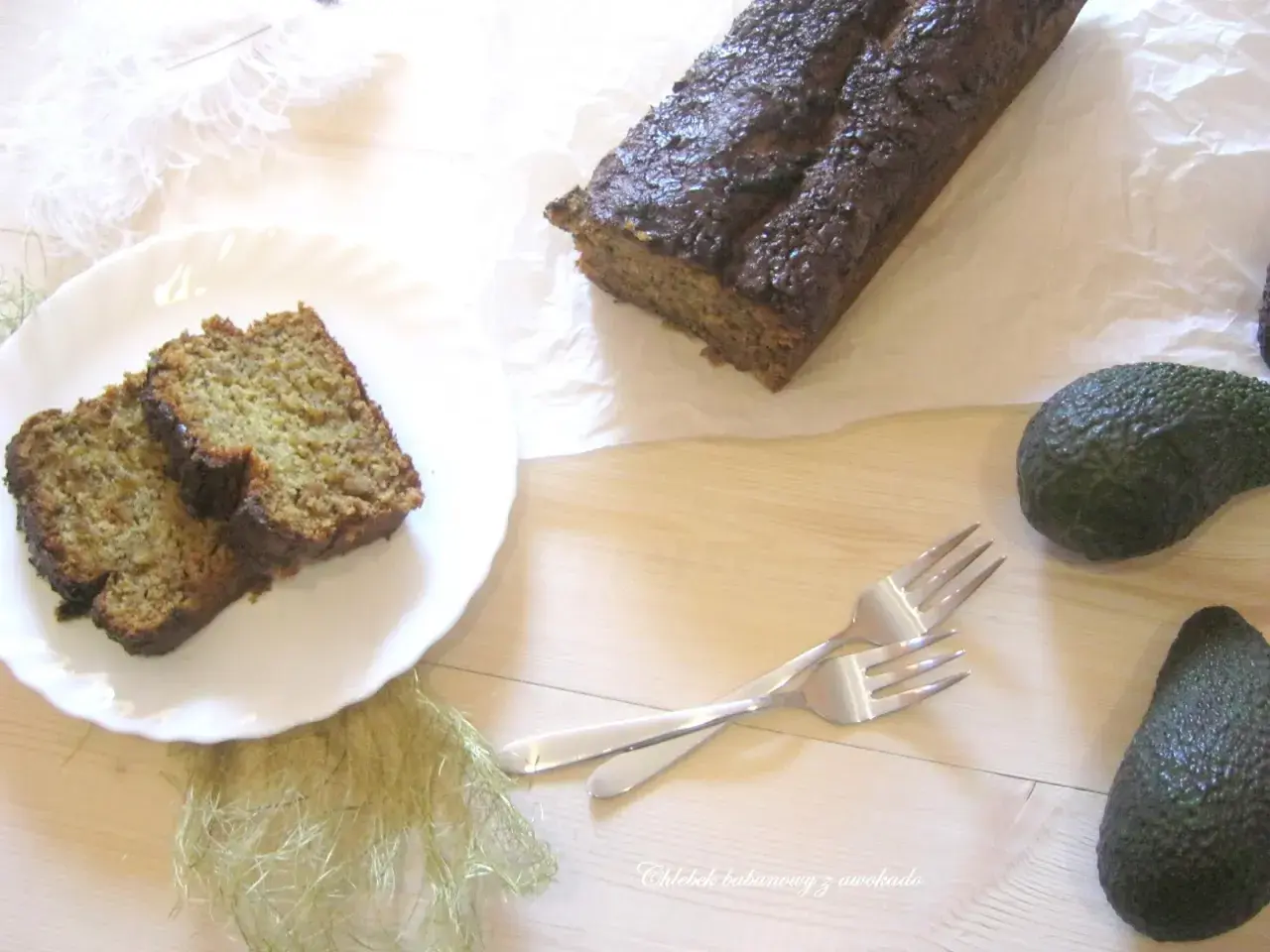 Chleb bananowy z awokado, pokrojony na kromki, leży na talerzu obok całego bochenka. Dwa awokado i widelce dopełniają kompozycję.
