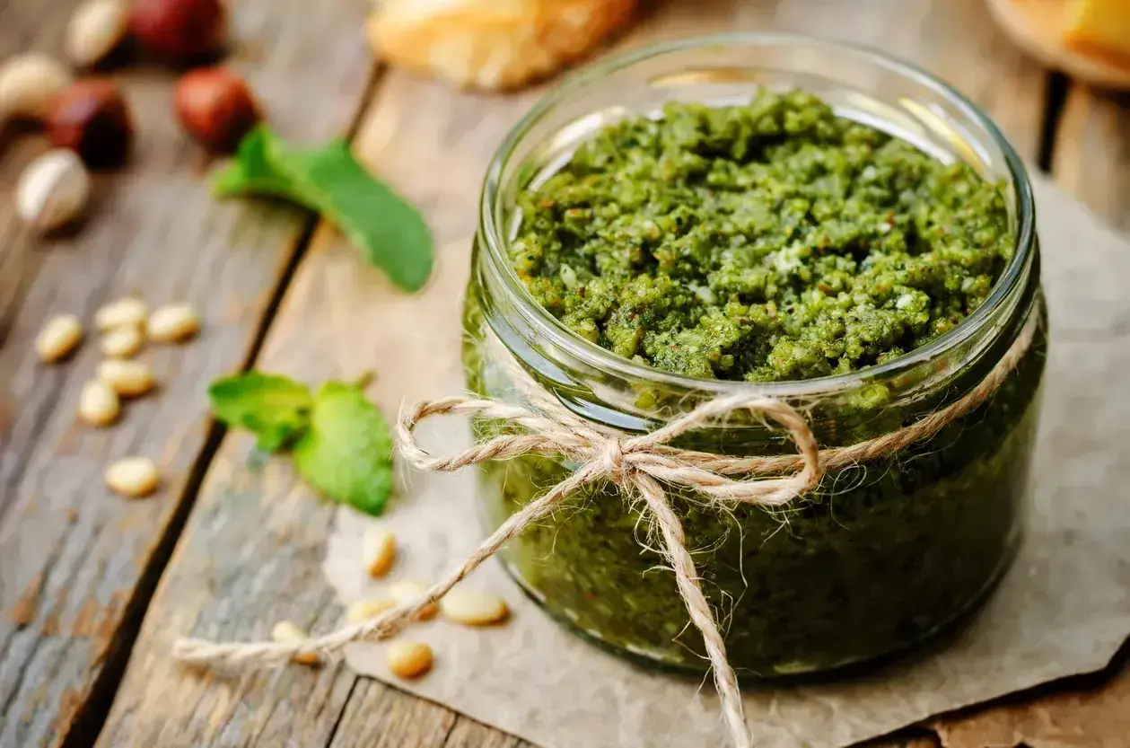 Słoik domowego pesto bazyliowego, ozdobiony sznurkiem. Obok leżą orzeszki piniowe i listki bazylii.