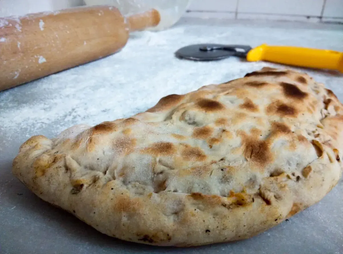 Złocisty calzone, gotowy do podania. Obok wałek i nóż do pizzy, jakby ktoś właśnie skończył przygotowywać ten pyszny przepis.